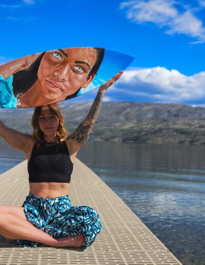 Amber Bahm holding up realistcc portrait painting on surfboard in front of lake in Peachland, BC